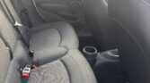 The image shows the back seat area of a mini car with black fabric seats, seat belts, and a cup holder between the front seats. The car door is closed, and outside, a wooden fence and houses are visible through the window.