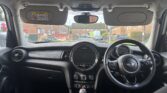 View from inside a mini car, showing the dashboard, steering wheel, and manual gear stick. Sun visors are down and outside, brick houses, parked cars, and a grassy area can be seen through the windshield on a quiet residential street.