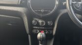 The photo shows the interior front view of a Mini car, focusing on the center console with a circular display, air vents, climate controls, toggles, a manual gear stick with stitching, and partial views of the front seats and steering wheel.