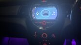A mini car interior at night shows a circular digital dashboard with a blue-lit screen displaying navigation and music icons. Red and purple lights illuminate the controls as an air freshener hangs above, with houses visible through the window.