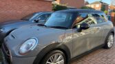 A silver Mini car with a black roof is parked on a brick driveway beside another silver car, in front of a red brick house with a tiled roof. The Mini has water droplets on its surface, indicating it was recently wet.