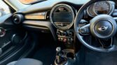 A photo of a Mini car interior showing the dashboard, steering wheel with Mini logo, touchscreen display, manual gearshift, air vents, and climate controls. The black seats and sunlight brighten the cabin, with a brick wall visible outside.