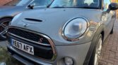 A close-up view of a grey Mini Cooper S, a classic mini car with raindrops on its surface, parked on a brick driveway next to another car. The license plate reads "YE67 AFX" and sunlight reflects off the hood and windshield.