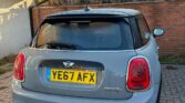 A grey Mini Cooper S, a stylish mini car with a black roof and twin exhausts, is parked on a brick driveway before a red brick wall. The car's UK plate reads "YE67 AFX," with bushes and part of a house in the background under clear skies.