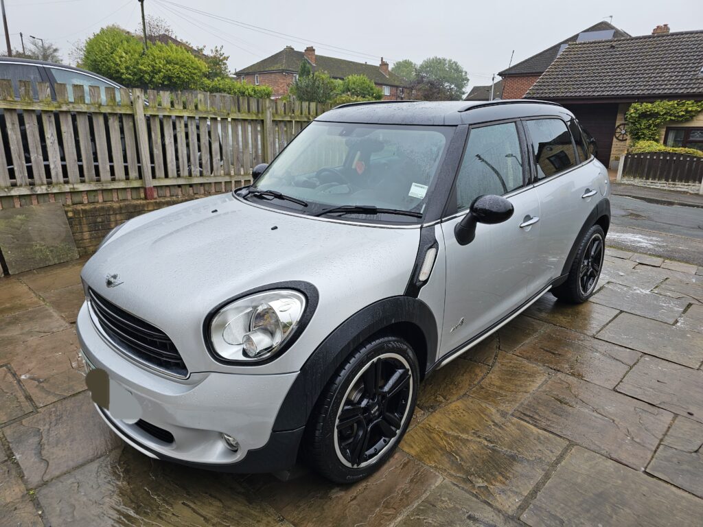 A silver Mini Countryman is parked on a wet stone driveway in front of a wooden fence and houses. This stylish mini car features black wheel rims and roof, with raindrops glistening on its surface under an overcast, rainy sky.