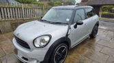 A silver Mini Countryman is parked on a wet stone driveway in front of a wooden fence and houses. This stylish mini car features black wheel rims and roof, with raindrops glistening on its surface under an overcast, rainy sky.