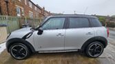 A silver mini car with black roof and trim is parked on a wet stone driveway in front of a wooden fence, brick houses, and a cloudy sky. The Mini is viewed from the side, showing four doors and striking black alloy wheels.