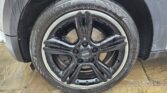 Close-up of a black alloy wheel with five double spokes on a MINI car. The tire is labeled "Ecsta" and is slightly wet, suggesting recent rain. Parked on tan stone paving, part of the mini trader’s grey body is visible.