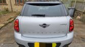 A rear view of a silver MINI Countryman car parked on a stone driveway in front of a house, perfect for any mini trader. The car has tinted rear windows, distinct MINI and Countryman badges, and a partially obscured license plate.