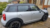 A silver mini car Countryman with black wheels is parked on a wet stone driveway in front of a tan brick house with wooden trim. Bushes and another house sit in the background, and rain speckles the mini’s windows.