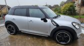 A silver Mini Countryman car with black accents and wheels is parked on a wet stone driveway in a residential area. This stylish mini car, viewed from the side, highlights its four doors, compact design, houses, greenery, and an overcast sky.