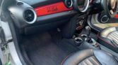 The image shows the front interior of a mini car, specifically the driver's side. The dashboard features a bright red panel with a signature, round air vents, black-and-red leather seats, and a center console with cup holders and control dials.