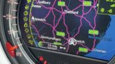 A close-up of a Mini car dashboard displays a GPS navigation screen showing the Ipswich area, including Thetford, Sudbury, and Saxmundham. The mini trader’s screen shows a 10 mph speed limit, time as 8:23, and temperature at -6°C.