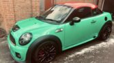 A bright green mini car coupe with a red roof and black wheels is parked on a driveway next to a brick wall. The sporty Mini stands out with tinted windows, red brake calipers, and its bold color combination—a true head-turner for any mini trader.