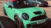 A matte green Mini Cooper coupe with a red roof is parked in a sunny outdoor lot. This sporty mini car features black wheels, red brake calipers, and a vented hood. Another gray car is nearby, with trees and a fence in the background.