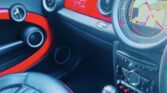 The photo shows the interior of a MINI car with black and red accents. The dashboard displays a large round GPS screen with a map. The seat is black leather, and several round air vents, control buttons, and the gear shift are visible in the foreground.