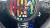 A close-up view of a MINI car steering wheel, featuring rainbow stripes and the MINI logo at the center. The dashboard is visible in the background, and a person wearing bright blue pants is seated behind the wheel—a perfect shot for any mini trader.