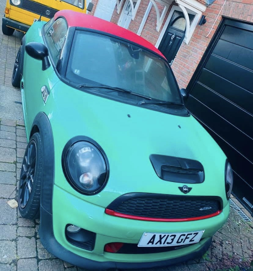 A lime green mini car with a red roof and black detailing is parked on a brick driveway in front of a house. The license plate reads "AX13 GFZ." A yellow van is visible in the background. Ideal for any mini trader enthusiast.