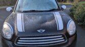 A black Mini car with white racing stripes is parked facing forward on a paved road near greenery and leafless trees. The UK license plate reads “MW11 XPC.” Some condensation is visible on the windshield of this stylish mini.