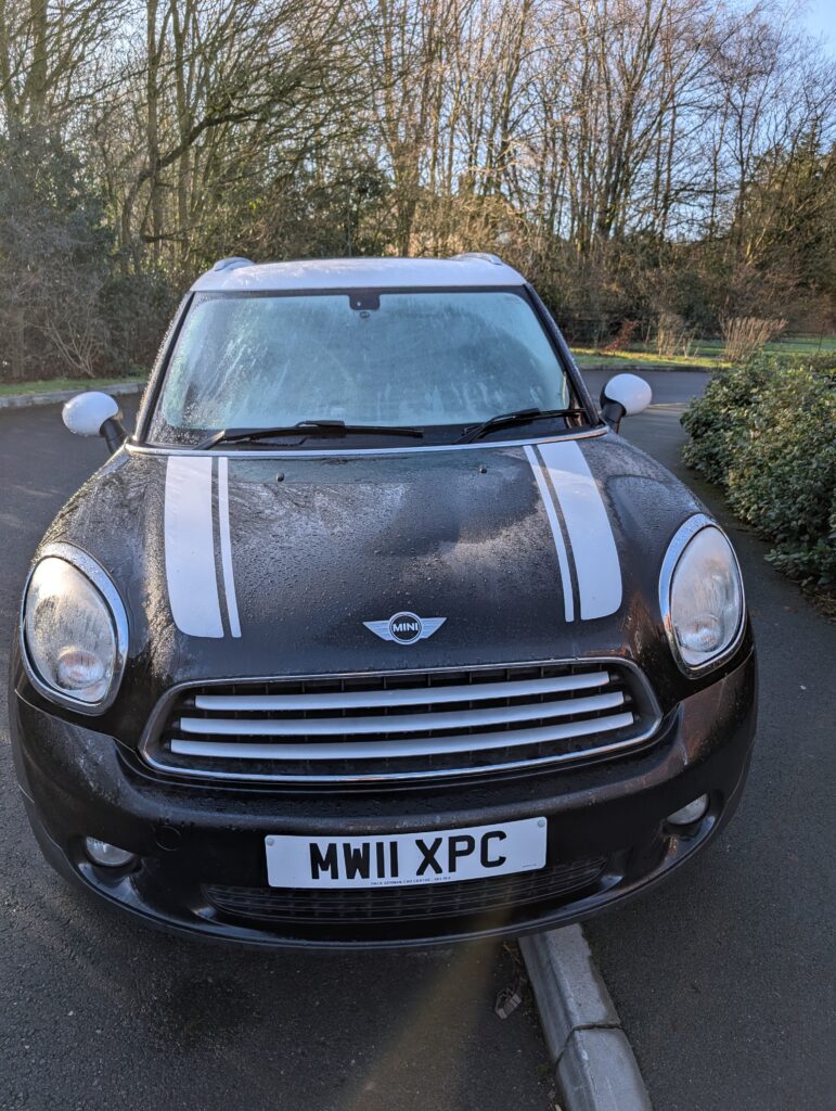 A black Mini car with white racing stripes is parked facing forward on a paved road near greenery and leafless trees. The UK license plate reads “MW11 XPC.” Some condensation is visible on the windshield of this stylish mini.