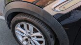 A close-up of a black Mini car’s front left tire parked on the street. The tire is flat, and droplets of water are visible on the car’s body, with reflections of nearby houses appearing on the wet surface in daylight.