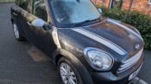 A black Mini Cooper Countryman—this stylish mini car with white racing stripes and a frosted windshield—is parked on a residential street near a brick house and green bushes. It features silver side mirrors and alloy wheels.