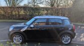 A black Mini Countryman mini car is parked on a paved road beside a hedge. Sunlight gleams on its surface, while leafless trees and blue sky form the backdrop, with patches of greenery at the bottom of the image.