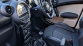 The photo shows the interior of a mini car, featuring the front seats and dashboard. The driver’s seat, steering wheel, manual gear stick, cup holders, and frosted windows are visible as sunlight highlights the black and beige upholstery.
