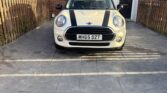 A classic white Mini Cooper with black racing stripes is parked on a driveway in front of a red brick house. If you’re searching for your perfect mini car, this scene could be your next driveway dream.