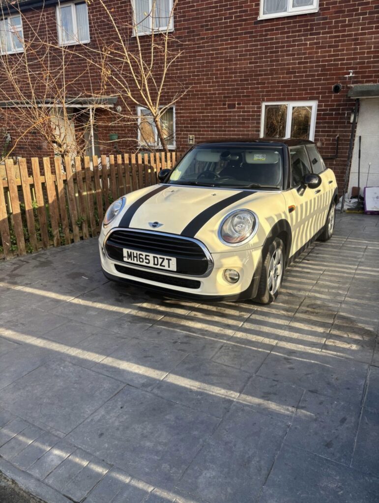 A cream-colored Mini Cooper, listed on Mini Trader, with black racing stripes is parked on a paved driveway beside a wooden fence in front of a brick house with white-framed windows. Leafless branches are visible by the fence.