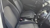 The image shows the interior of a mini car, focusing on the front passenger and driver seats covered in dark fabric. The dashboard, steering wheel, manual gear stick, and door panels are visible as sunlight brightens the compact space.