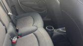 The image shows the backseat area of a mini car with dark gray fabric seats, a center armrest with cup holders, and seat belts fastened. The clean interior features a visible door and window, with houses and a wooden fence seen outside.