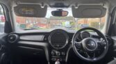 View from the back seat of a mini car, showing the dashboard, steering wheel with Mini logo, central display, rearview mirror, and two sun visors down. Outside, a suburban street with brick houses and parked cars is visible.