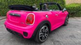 A bright pink mini car convertible is parked on asphalt near a hedge and brick building, its black soft top down. The sporty Mini features alloy wheels, black detailing, red taillights, and an empty rear license plate—perfect for a Mini trader.