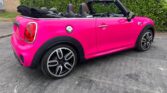 A bright pink Mini convertible with its black soft top down is parked on a paved area near a hedge and brick building. This stylish mini car features silver wheels, black accents, and visible front and rear seats with headrests.
