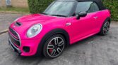 A bright pink mini car convertible with black accents and a black soft top is parked on asphalt beside a tall green hedge and a brick building. The mini features sporty black and silver wheels and red brake calipers.
