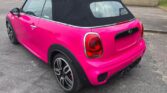 A bright pink Mini Cooper convertible—a stylish mini car with a black soft top—is parked on a paved surface near a low brick building. The sporty wheels, black trim, and lack of license plate highlight its unique look.