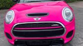 A bright pink mini car is parked on a dark asphalt lot, shown from the front with its headlights, grille, and logo visible. In the background, a green hedge and a brick and metal building with notices complete the scene.