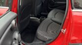 The image shows the rear passenger area of a red mini car with the door open. The interior features black fabric seats, visible seat belts, and a clean floor. The armrest and cup holder are between the seats. Part of the front seat and door are visible.