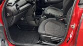The image shows the front interior of a red Mini car with the driver’s door open. The seats are black, the dashboard features a circular design, and there is a manual gear stick. The car is parked on a road beside another black vehicle.