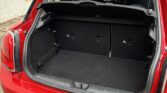 The image shows the open trunk of a red mini car, revealing an empty, clean, and carpeted cargo space. The rear seats are upright, with a black removable parcel shelf above the trunk area. The car is parked on a paved surface beside a light-colored wall.