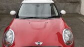 A red mini car with a white roof is parked facing forward in front of a stone wall. A blue "PRIVATE CAR PARKING" sign hangs nearby, while old stone buildings and the license plate "SM64 XUR" complete the classic Mini scene.
