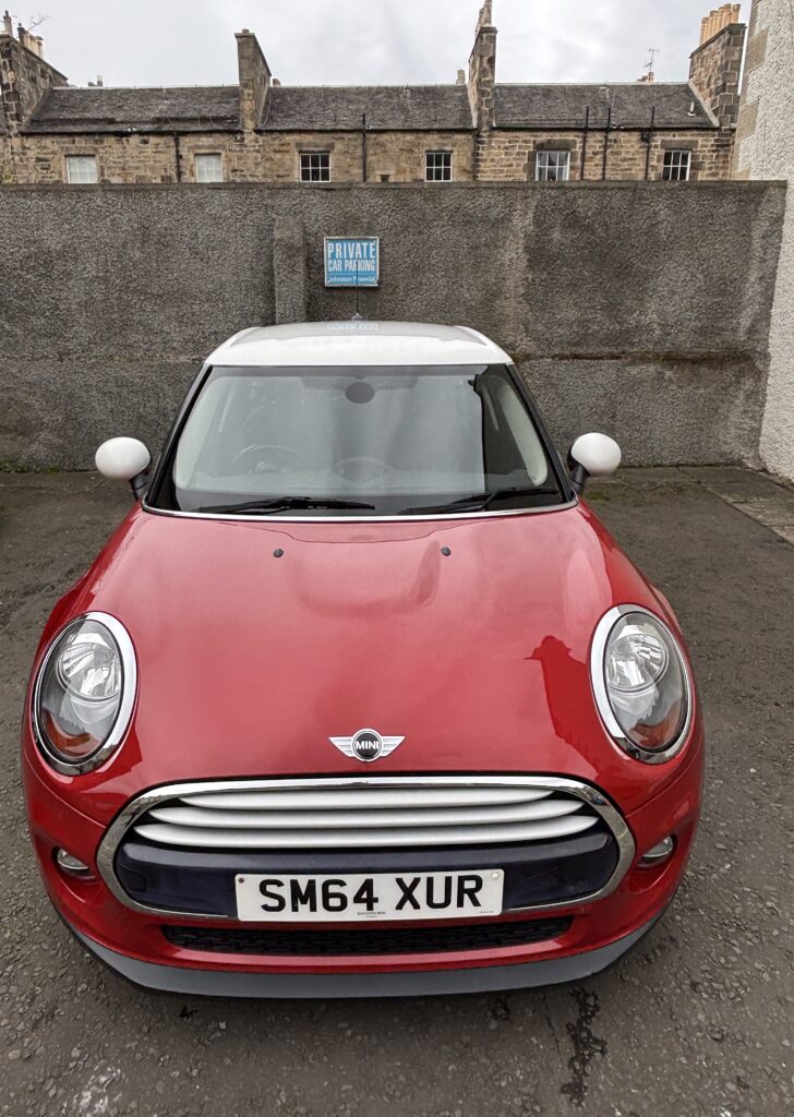 A red mini car with a white roof is parked facing forward in front of a stone wall. A blue "PRIVATE CAR PARKING" sign hangs nearby, while old stone buildings and the license plate "SM64 XUR" complete the classic Mini scene.