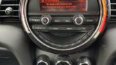A close-up view of a Mini car dashboard shows a round digital display playing BBC Radio 2 with "Stargazing by Myles Smith" now playing. Below are control buttons, three dials for climate settings, and toggle switches. The interior is black.