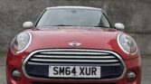 A red Mini Cooper mini car with a white roof, photographed from the front, is parked on a concrete surface with a stone wall and buildings in the background. The license plate reads "SM64 XUR" and the sky is overcast.