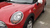 A red Mini car with a white roof and white side mirrors is parked in a lot beside a gray wall. A blue sign on the wall reads "PRIVATE CAR PARKING." Another black car is partially visible on the left. The ground is paved and slightly worn.