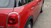 A classic red mini car with a white roof is parked on a cobblestone street near old stone buildings. The car's rear and left side are visible, showcasing its distinctive taillights and shiny surface. A green bin stands near a black door in the background.