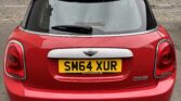 A red mini car with a white roof is parked on a cobblestone street. The rear view shows a yellow UK license plate reading "SM64 XUR." Stone buildings and a stone wall with foliage form the background.