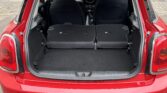 A red mini car hatchback with its rear hatch open, showing the spacious trunk area. The back seats are folded down flat, extending the cargo space. The car is parked on a paved surface with another car and a brown bin in the background.