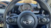 A person’s view from the driver’s seat of a mini car shows a black steering wheel with the Mini logo, dashboard gauges, and blue sky outside the windshield. The person’s legs, wearing blue pants, are visible beneath the wheel—perfect for any mini trader.
